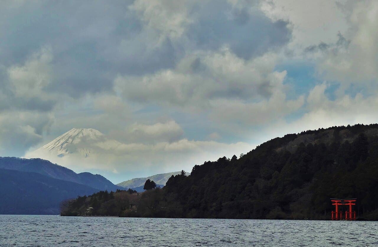 Lake Ashi with the red torii of Hakone Shrine at the wooded shore and snow-capped Mount Fuji partly veiled in cloud.